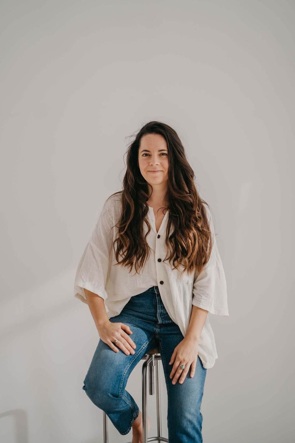 Angels Perez sitting on a stool in a cream shirt and jeans, looking at the camera